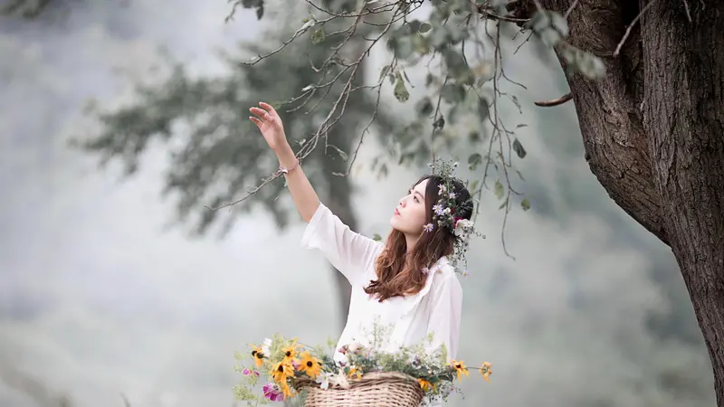 A woman in white with a basket of flowers reaching up to a branch in soft mist