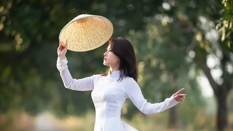 A woman in a flowing white ao dai holding a hat on a tree-lined road