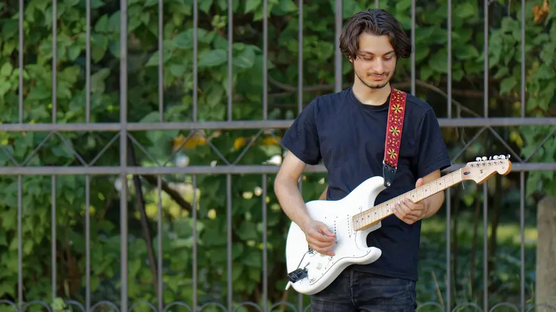 A young man playing a white electric guitar in front of a green fence